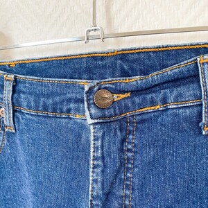 May include: Close-up of blue denim jeans with orange stitching. The jeans feature a button closure and a metal button with a logo. The jeans are hanging on a metal hanger against a white background.
