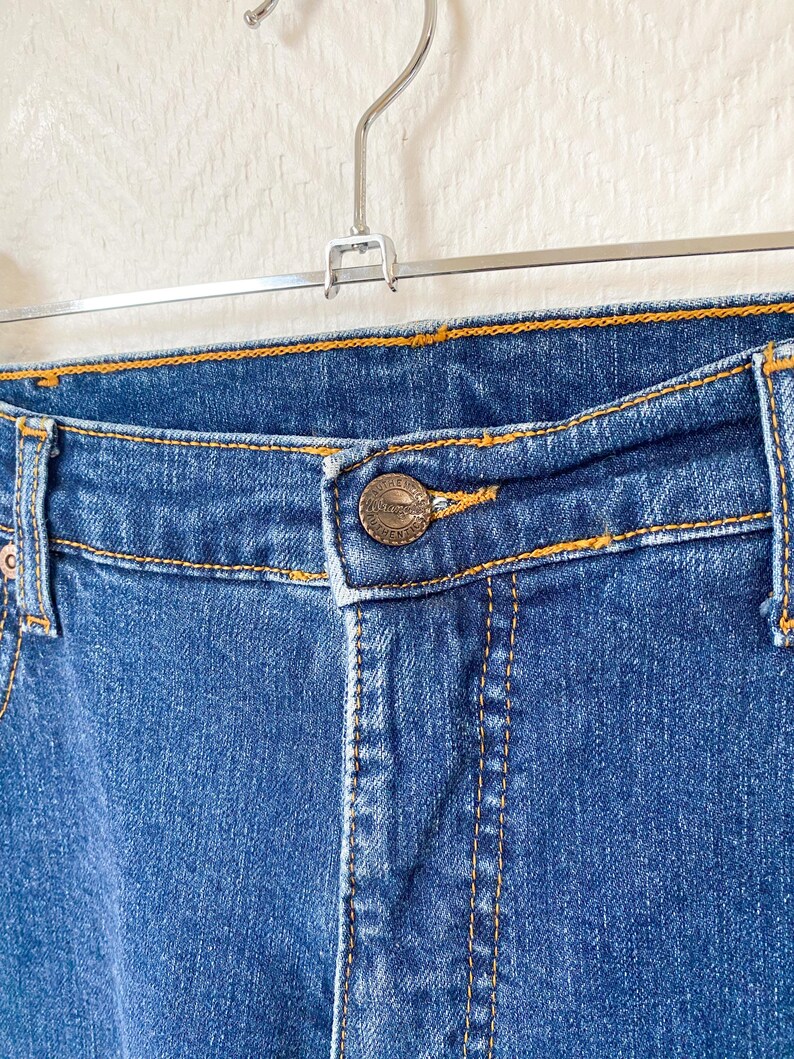 May include: Close-up of blue denim jeans with orange stitching. The jeans feature a button closure and a metal button with a logo. The jeans are hanging on a metal hanger against a white background.