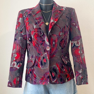 Può includere: Un blazer fantasia nei toni del grigio, rosso e viola, esposto su un manichino. Il blazer presenta una chiusura con un solo bottone e un design floreale. Il manichino indossa anche una collana d'oro e pantaloncini di jeans.