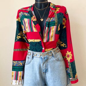 Peut inclure: Une chemise boutonnée à motifs rouges, verts et dorés à manches longues, associée à un short en jean bleu clair et une ceinture chaîne dorée. La chemise présente un motif floral et géométrique. Un buste noir présente la tenue.
