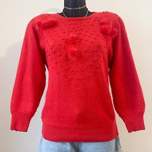 Può includere: Un maglione rosso acceso con maniche lunghe e scollo tondo. Il maglione è decorato con piccole perline rosse e tre abbellimenti strutturati e soffici. L'indumento è esposto su un manichino nero, abbinato a pantaloncini di jeans.