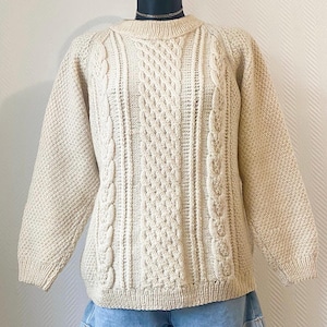 Peut inclure: Pull-over en tricot crème avec un col rond et des manches longues. Le pull présente des motifs de câbles complexes sur le devant et le long des manches, avec un motif de point texturé. Le vêtement est présenté sur un mannequin.