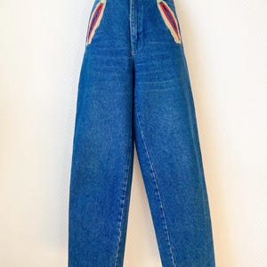 Op de afbeelding: Een high-waisted, wijd uitlopende blauwe jeans. De jeans heeft decoratieve rode en crèmekleurige accenten op de voorzakken en tailleband. De jeans heeft een knoop- en ritssluiting en is gemaakt van een medium-wash denim.