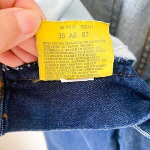 May include: Close-up of a yellow tag with black text attached to blue denim fabric. The tag includes washing instructions and the numbers "56 09 01 1135441" and "39 AG 07". The denim is a deep indigo color.
