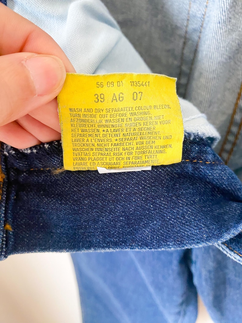 May include: Close-up of a yellow tag with black text attached to blue denim fabric. The tag includes washing instructions and the numbers "56 09 01 1135441" and "39 AG 07". The denim is a deep indigo color.