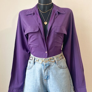 Könnte beinhalten: Ein lila Hemd mit Knöpfen und zwei Brusttaschen, kombiniert mit hellblauen Jeansshorts und einem goldfarbenen Ketten-Gürtel. Das Outfit wird durch mehrlagige goldfarbene Halsketten, einschließlich eines Herzanhängers, ergänzt. Kleidung auf einer schwarzen Schaufensterpuppe.