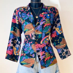 Puede incluir: Una blazer vintage con un vibrante estampado floral en tonos rosa, morado, verde y marrón sobre un fondo negro. La blazer tiene mangas largas, cuello con solapas y botones de madera. Se lleva con pantalones cortos vaqueros.