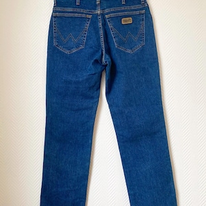 May include: Blue denim jeans with classic five-pocket styling. Features include a brown leather patch and signature stitching on the back pockets. The jeans are hung on a metal hanger.
