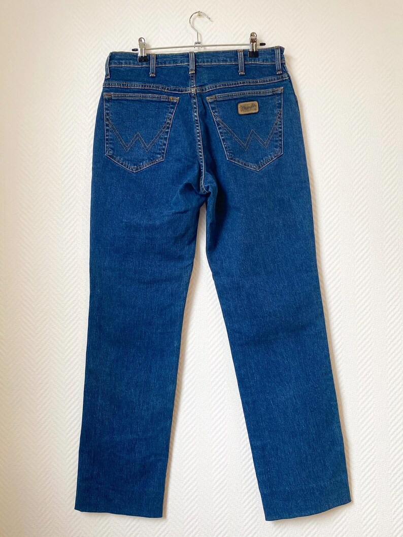 May include: Blue denim jeans with classic five-pocket styling. Features include a brown leather patch and signature stitching on the back pockets. The jeans are hung on a metal hanger.