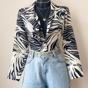 Pode incluir: Um blazer com estampa de zebra em preto e branco combinado com calções de ganga azul claro. O conjunto é complementado com um cinto de corrente dourada e colares dourados em camadas. Os calções têm bainhas dobradas.