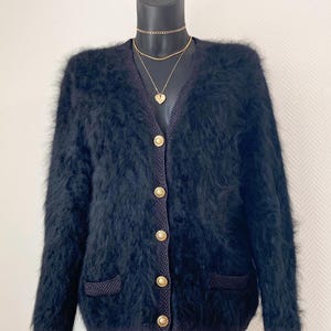 Peut inclure: Un cardigan noir à manches longues en maille duveteuse avec un col en V, cinq boutons circulaires dorés sur le devant et deux petites poches en bas. Le vêtement présente une finition en maille ébouriffée avec des bords côtelés.
