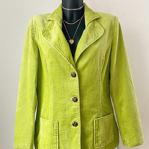 Op de afbeelding: Een limoengroene corduroy blazer met een reverskraag, drie bruine knopen en twee voorzakken. De blazer wordt gepresenteerd op een zwarte mannequin. Een gouden ketting is zichtbaar.
