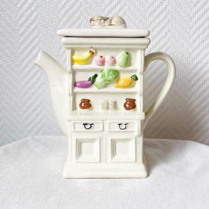 Peut inclure: Théière en céramique blanche en forme de meuble, ornée de fruits et légumes miniatures colorés sur des étagères. Une figurine de chat endormi repose sur le couvercle. La théière a un bec et une anse.