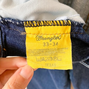 May include: Close-up of a yellow Wrangler tag on dark blue denim jeans. The tag displays the brand name, size 33-34, fabric composition, and the word "TEXAS". The jeans are partially visible.