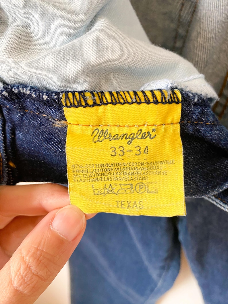 May include: Close-up of a yellow Wrangler tag on dark blue denim jeans. The tag displays the brand name, size 33-34, fabric composition, and the word "TEXAS". The jeans are partially visible.