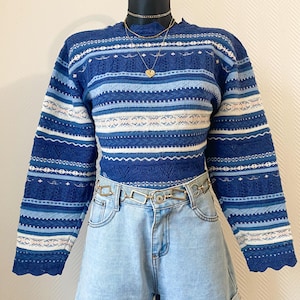 Peut inclure: Un pull à motifs bleus et blancs à manches longues, associé à un short en jean bleu clair et une ceinture chaîne dorée. Le pull a un col montant et un motif tricoté détaillé. Un collier doré avec un pendentif en forme de cœur complète la tenue.