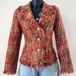 Peut inclure: Veste en tweed multicolore rouge, orange et vert, avec des franges sur le col, les poignets et l'ourlet. La veste a trois boutons argentés et est présentée sur un mannequin. Elle est associée à un short en jean.