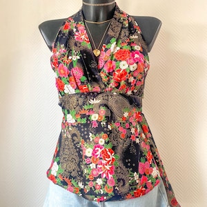Peut inclure: Un haut dos nu noir orné d'un motif floral vibrant avec des fleurs rouges, roses et blanches, accentué par des lignes dorées de style cachemire. Le haut présente un décolleté en V, une large ceinture et un ourlet évasé, porté avec un short en jean bleu clair.