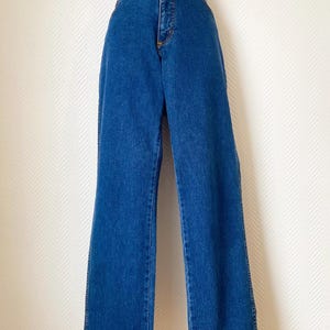 May include: Blue denim jeans with a high waist and wide leg. The jeans have a classic five-pocket design, a zipper fly, and a button closure. The denim is a medium wash, and the jeans appear to be in good condition.