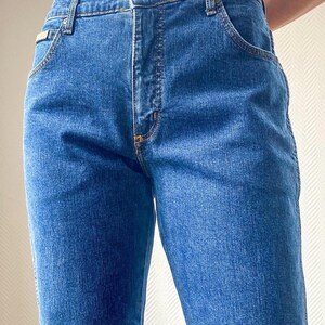 May include: Close-up of blue denim jeans. The jeans have a classic five-pocket design with a button and zipper closure. The stitching is a contrasting yellow color. The jeans are a medium wash and appear to be well-worn.