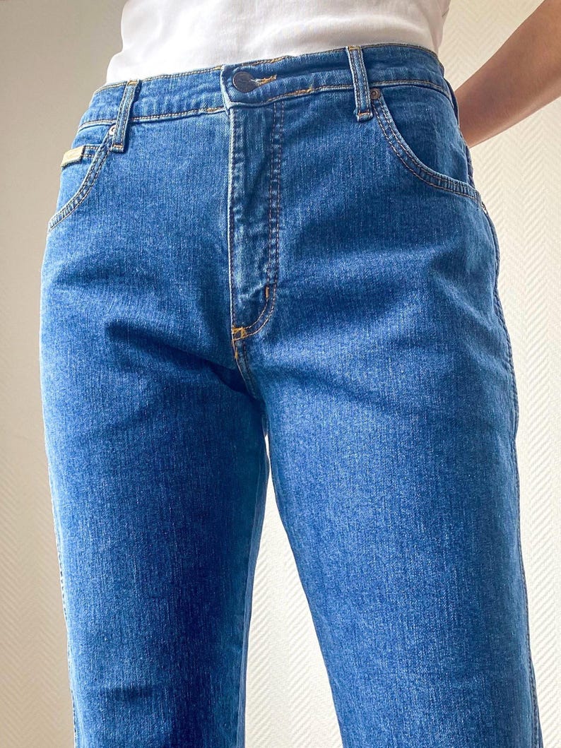 May include: Close-up of blue denim jeans. The jeans have a classic five-pocket design with a button and zipper closure. The stitching is a contrasting yellow color. The jeans are a medium wash and appear to be well-worn.