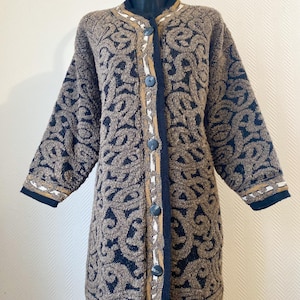 Peut inclure: Longue veste cardigan à boutons, ornée d'un motif marron et bleu marine. Le pull présente un motif texturé avec un motif tourbillonnant répété. Les bords sont ornés d'accents bleu marine, dorés et blancs. Le cardigan est présenté sur un mannequin.