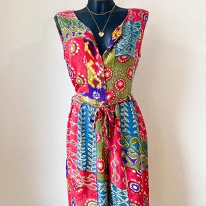 Op de afbeelding: Een mouwloze, knielange jurk met een patchwork ontwerp in rood, blauw, groen en goud. De jurk heeft een V-hals, knoopsluiting en een bijpassende strikceintuur. Een gouden ketting met een hartvormige hanger is zichtbaar.