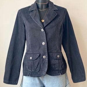 Peut inclure: Veste noire avec un design classique, col cranté, trois boutons argentés et deux poches avant à boutons. La veste est présentée sur un mannequin avec un collier doré et un pendentif en forme de cœur. Elle est associée à un short en jean.