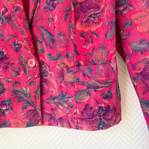 May include: A bright pink corduroy blazer featuring a dark floral pattern in shades of purple, orange, and green. The jacket includes a single front button closure, a notched lapel, and a horizontal flap pocket on the lower front section.