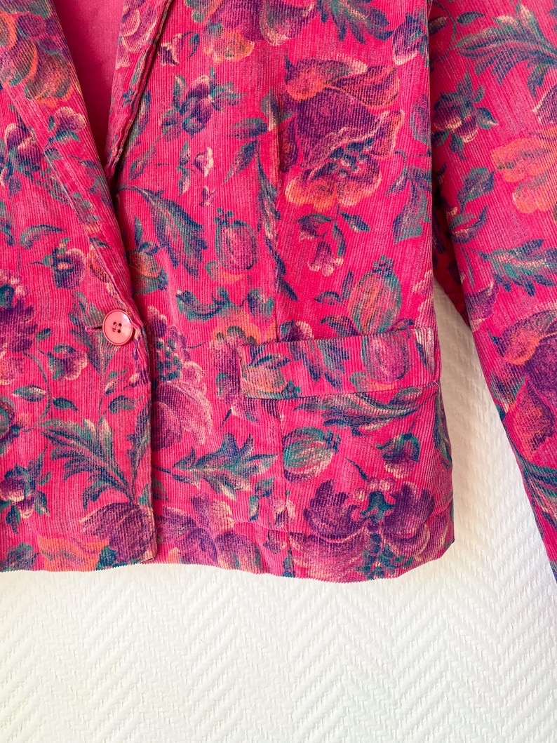 May include: A bright pink corduroy blazer featuring a dark floral pattern in shades of purple, orange, and green. The jacket includes a single front button closure, a notched lapel, and a horizontal flap pocket on the lower front section.
