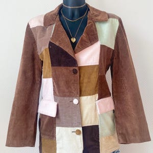 Peut inclure: Blazer en patchwork de velours côtelé dans les tons marron, rose et vert. La veste présente un col, deux poches avant et deux boutons. Le blazer est présenté sur un mannequin avec un collier doré.
