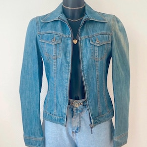 Könnte beinhalten: Hellblaue Jeansjacke mit Reißverschluss, zwei Brusttaschen und langen Ärmeln. Die Jacke wird mit hellblauen Jeansshorts und einer goldfarbenen Halskette mit Herzanhänger kombiniert. Ein goldfarbener Gürtel ist ebenfalls sichtbar.