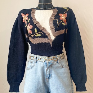Peut inclure: Un pull noir orné de broderies florales roses, oranges et vertes. Le pull présente un motif blanc et marron sur le devant. Il est associé à un short en jean bleu clair et une ceinture chaîne dorée.