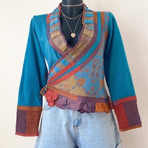 Peut inclure: Un haut portefeuille turquoise à manches longues avec une bordure à motifs colorés. Le haut présente un ourlet à volants et est associé à un short en jean. Les manches et la garniture ont des accents orange, violets et marron. Un collier en or est visible.