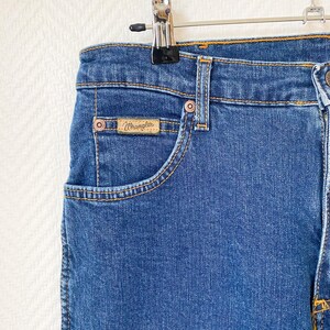 May include: Close-up of blue denim jeans with a classic five-pocket design. The jeans feature a small pocket with a "Wrangler" brand label. The jeans are hanging on a metal hanger.