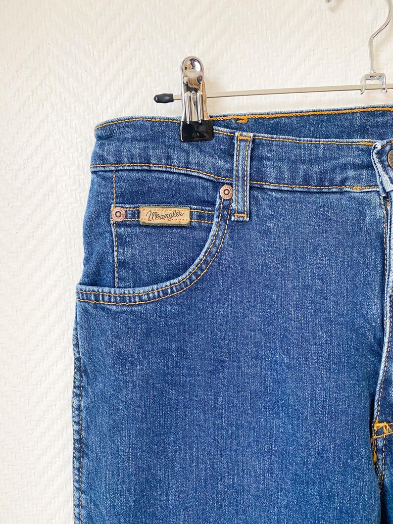 May include: Close-up of blue denim jeans with a classic five-pocket design. The jeans feature a small pocket with a "Wrangler" brand label. The jeans are hanging on a metal hanger.