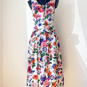 Peut inclure: Une robe midi sans manches avec une base blanche et un imprimé floral multicolore vif. La robe présente un corsage ajusté avec une fermeture boutonnée sur le devant, une taille marquée et une jupe ample froncée tombant sous les genoux.