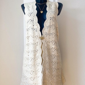 Puede incluir: Chaleco de crochet color crema, abierto por delante, con borde festoneado. El chaleco se exhibe en un maniquí y presenta un diseño calado delicado. Se ven un broche dorado y un collar con un colgante de corazón.