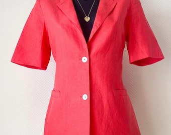 Pode incluir: Um blazer de manga curta num tom vermelho coral vibrante com lapela, dois botões brancos na parte central frontal e dois bolsos horizontais com debrum na cintura. A peça é exibida num manequim preto com um colar de corrente dourado.