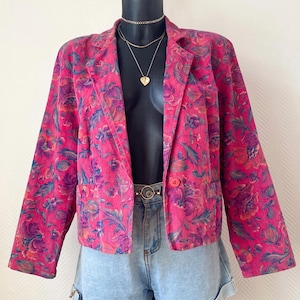 May include: A bright pink corduroy blazer featuring a vibrant floral pattern in shades of purple, blue, and orange. The jacket has long sleeves, a notched lapel, and a single button closure at the front. It is paired with light blue denim shorts.