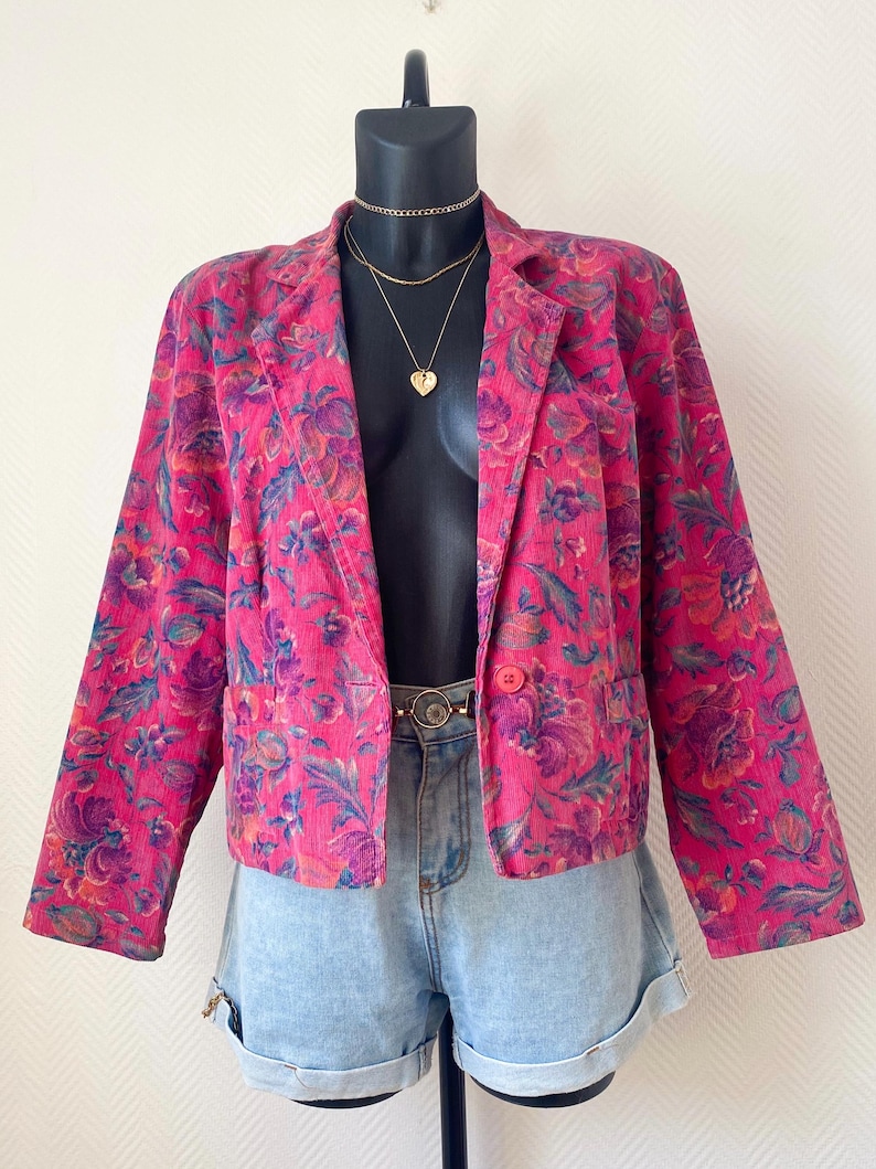 May include: A bright pink corduroy blazer featuring a vibrant floral pattern in shades of purple, blue, and orange. The jacket has long sleeves, a notched lapel, and a single button closure at the front. It is paired with light blue denim shorts.