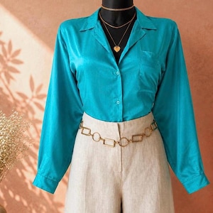 May include: A bright turquoise long-sleeve button-down shirt with a collar and a single chest pocket, paired with beige trousers and a gold-toned chain belt featuring rectangular and circular links, worn with a gold heart-shaped pendant necklace.