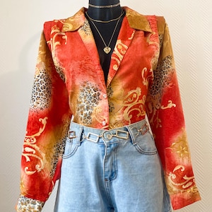 Peut inclure: Une chemise boutonnée aux motifs rouge, orange et or, avec des manches à imprimé léopard. Elle est associée à un short en jean bleu clair et une ceinture dorée, présentée sur un mannequin noir. Le motif de la chemise est de type cachemire.