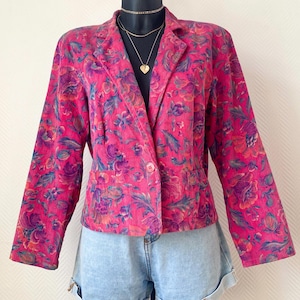 May include: A bright pink corduroy blazer featuring a vibrant floral pattern in shades of purple, orange, and teal. The jacket has a notched lapel, long sleeves, and a single front button closure. It is paired with light blue denim shorts.