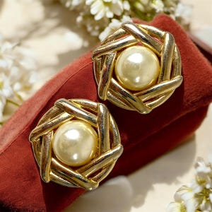 Peut inclure: Paire de boucles d'oreilles dorées à clip, avec un motif tressé entourant une grosse fausse perle crème. Les boucles d'oreilles sont posées sur un tissu de velours rouge foncé, avec des fleurs blanches en arrière-plan.