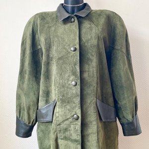 Puede incluir: Chaqueta de ante verde oliva con cuello, puños y detalles de bolsillo de cuero gris oscuro. La chaqueta tiene cierre de botones y dos bolsillos delanteros. La chaqueta se exhibe en un maniquí negro.