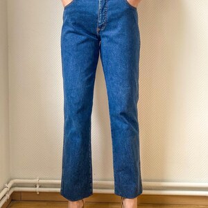 May include: Blue denim jeans with a classic five-pocket design. The jeans have a straight leg cut and a medium wash. The model is wearing gold-colored high-heeled sandals. The jeans are a vintage style.