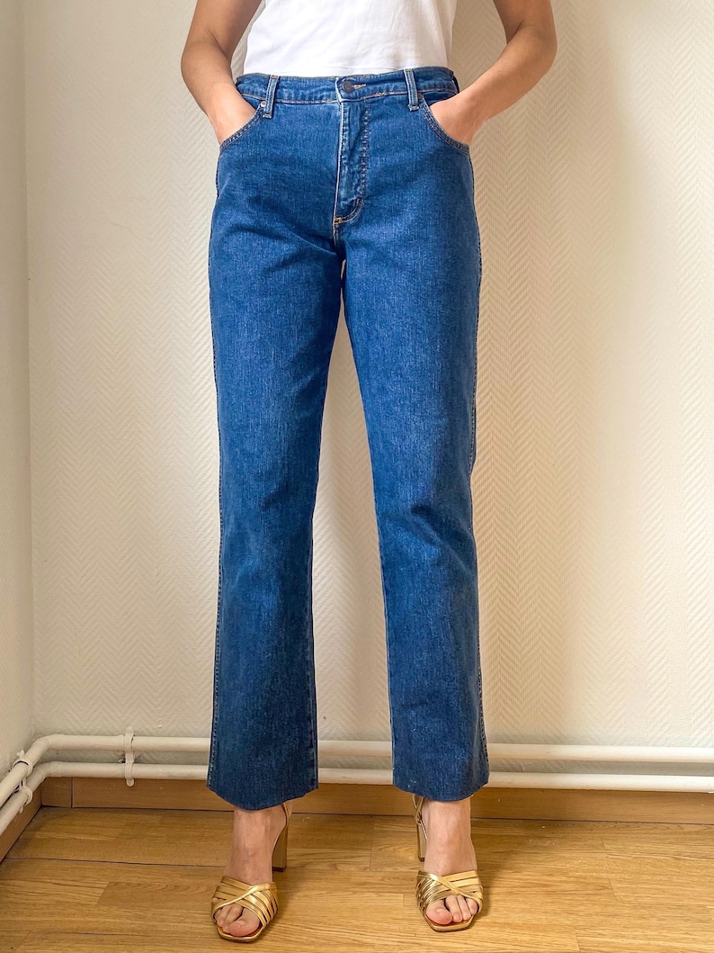 May include: Blue denim jeans with a classic five-pocket design. The jeans have a straight leg cut and a medium wash. The model is wearing gold-colored high-heeled sandals. The jeans are a vintage style.