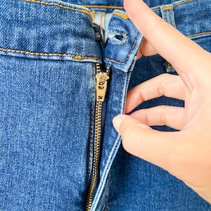 May include: Close-up of blue denim jeans with a gold zipper. The zipper is partially open, and the fabric shows detailed stitching. The jeans are a medium wash, and the image highlights the texture and construction of the garment.