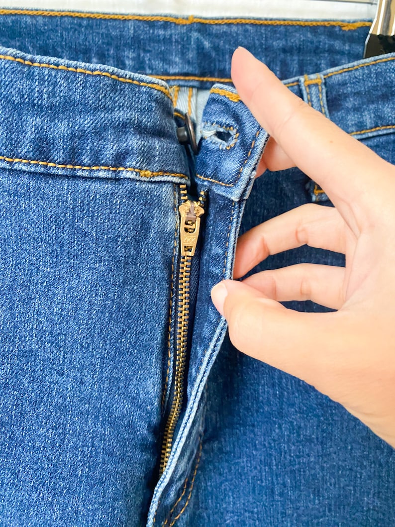 May include: Close-up of blue denim jeans with a gold zipper. The zipper is partially open, and the fabric shows detailed stitching. The jeans are a medium wash, and the image highlights the texture and construction of the garment.